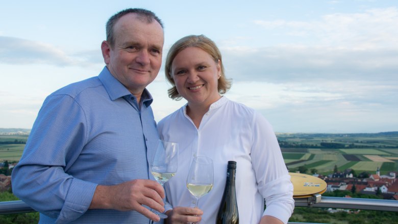 A man and a woman are holding wine glasses and smiling at the camera, with a landscape of fields in the background.