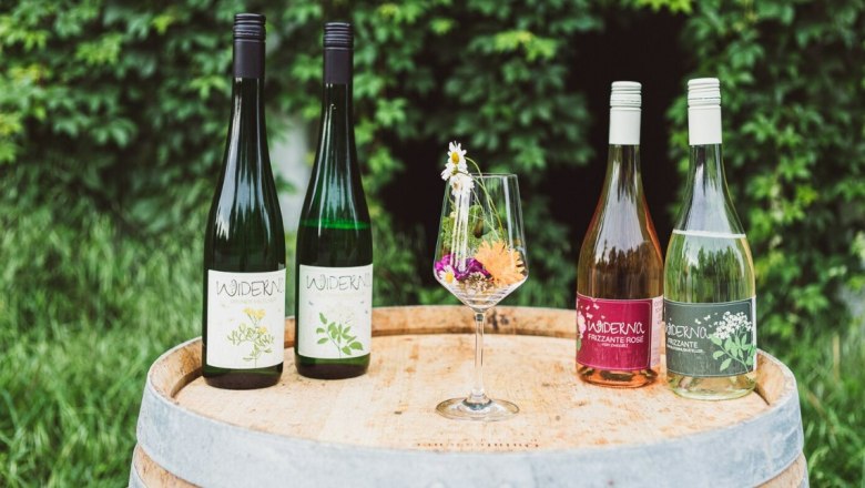 Four wine bottles and a wine glass with flowers on a wooden barrel in front of a green hedge.
