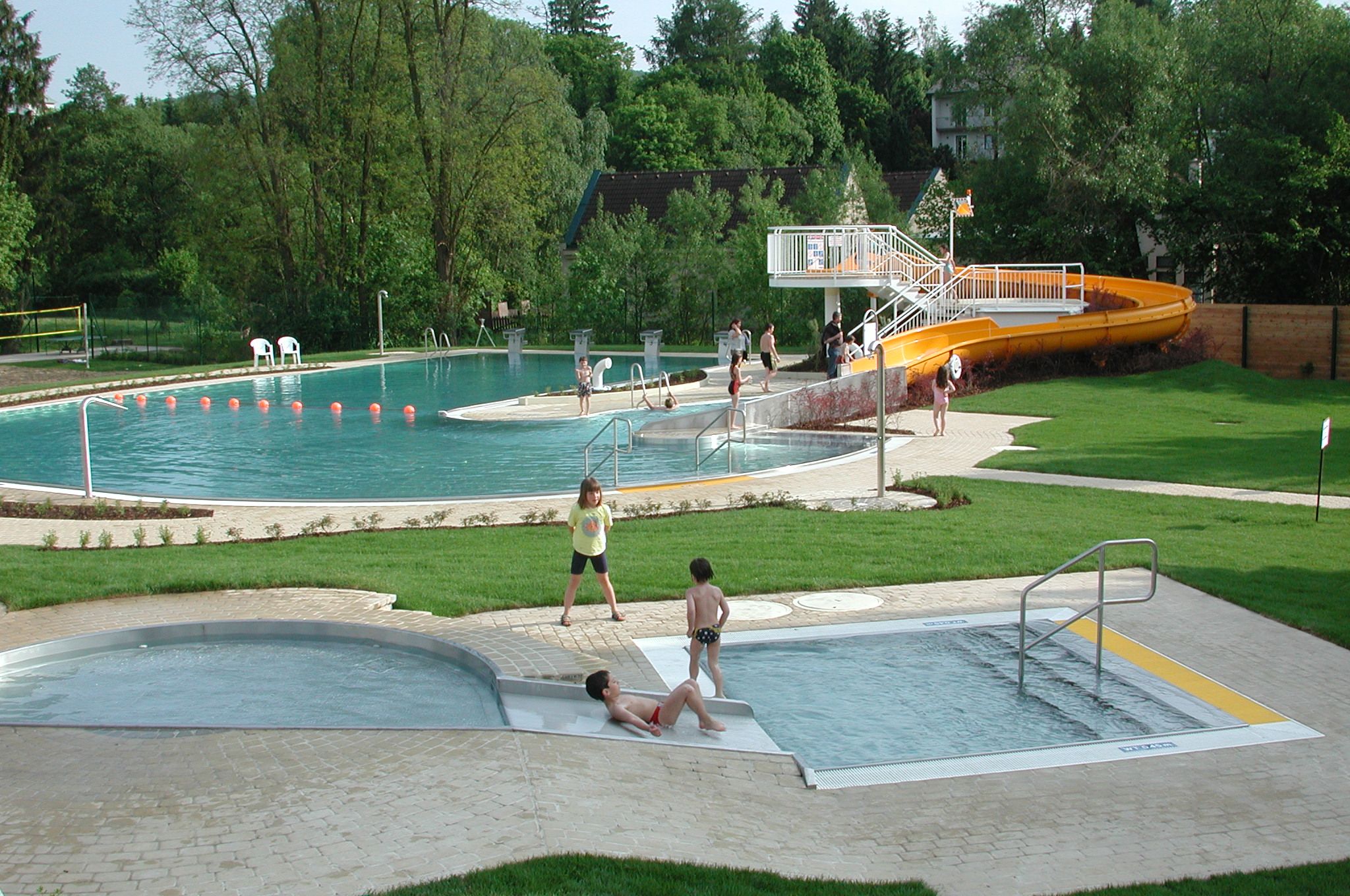 Outdoor pool with children's pool and slide, surrounded by trees and lawns.