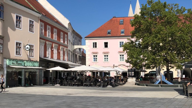 A square with cafés and stores, parasols and a tree.