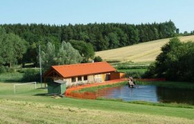 A small bathing pond with a wooden building and surrounding fields and woods.
