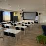An empty seminar room with white tables, black chairs and a large screen at the front.