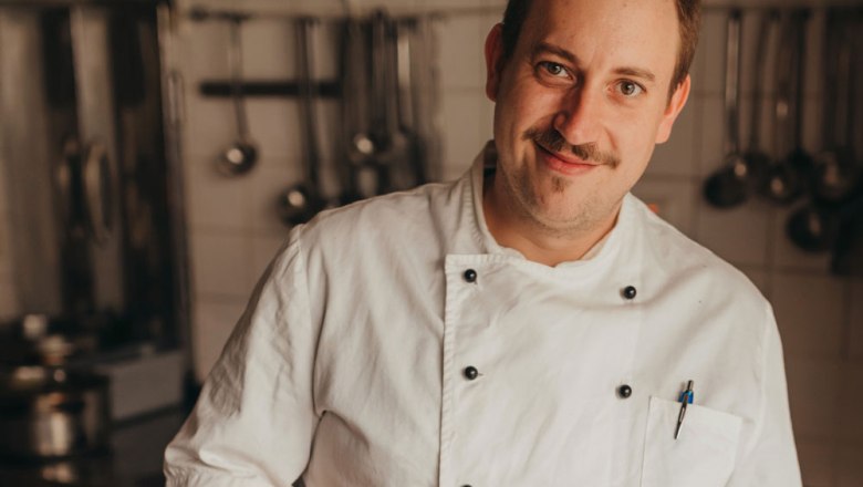 A chef in a white uniform smiles at the camera, a kitchen with hanging ladles in the background.