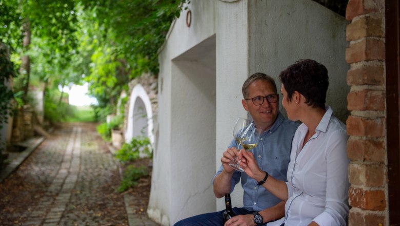 A couple sits in a wine cellar lane and toasts with glasses of wine.