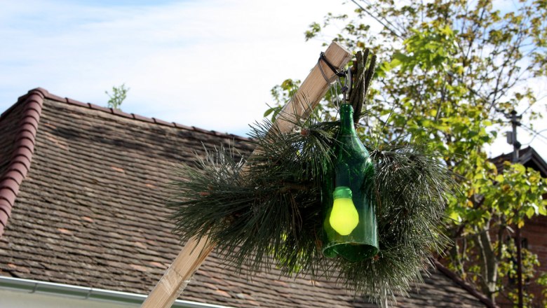 A green bottle with a yellow light bulb hangs on a wooden pole, surrounded by fir branches, in front of a roof and trees.