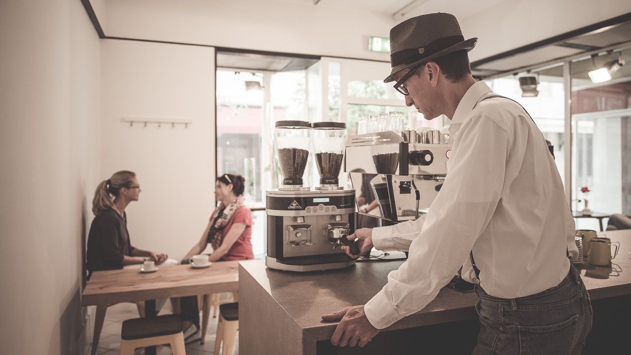 A barista prepares coffee in a stylish café while two women sit at a table chatting.