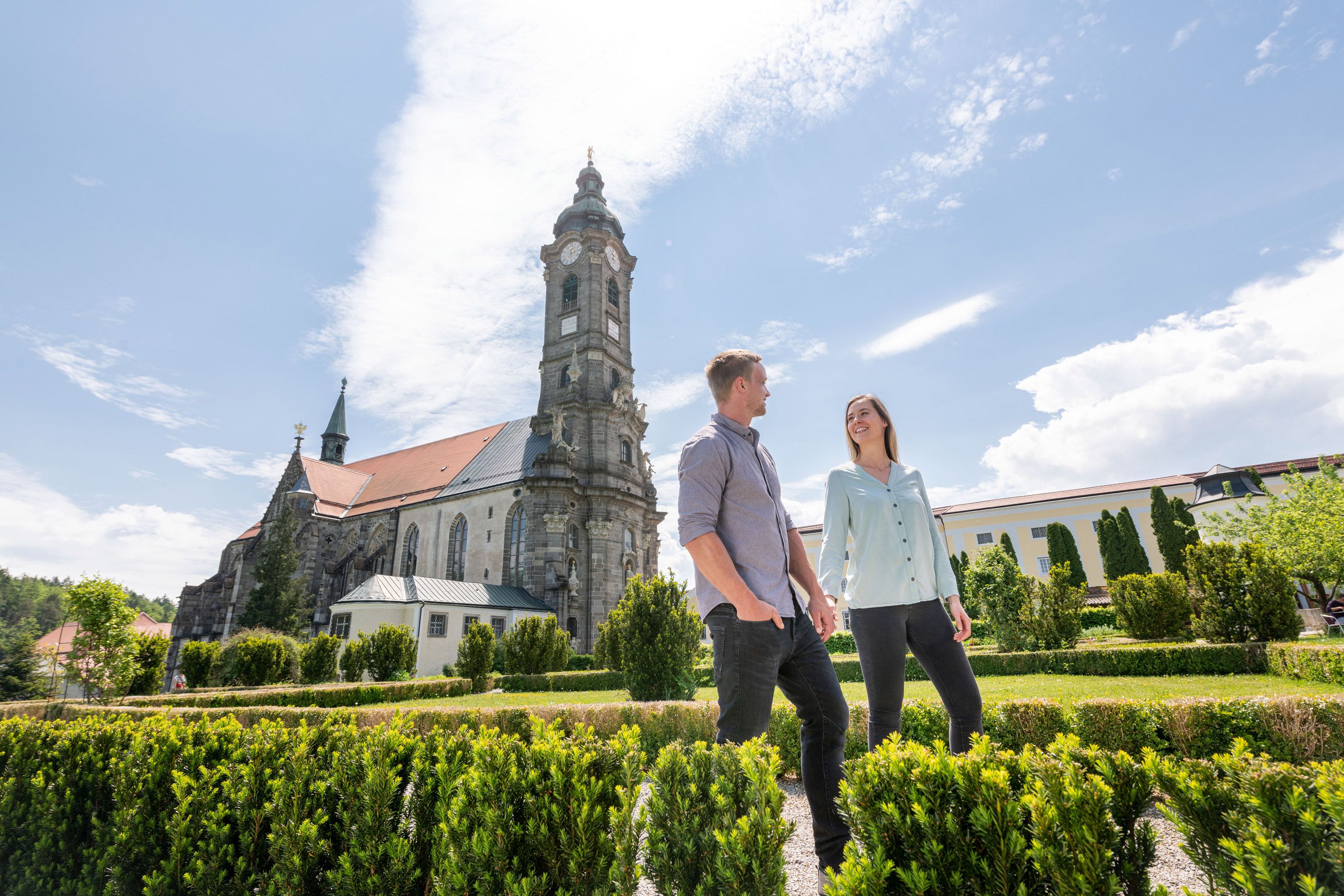 A couple walks in the garden of Zwettl Abbey, with the impressive architecture of the monastery in the background.