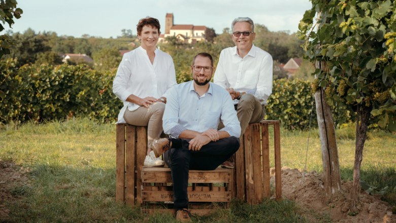 Three people sitting on wooden crates in a vineyard, with a church in the background.