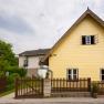 A yellow house with a pointed roof and wooden fence in a rural setting.