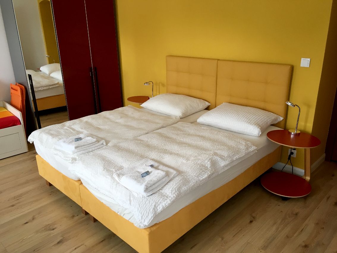 A double bed with a yellow headboard, white comforters and pillows in a room with a yellow wall and red wardrobe.