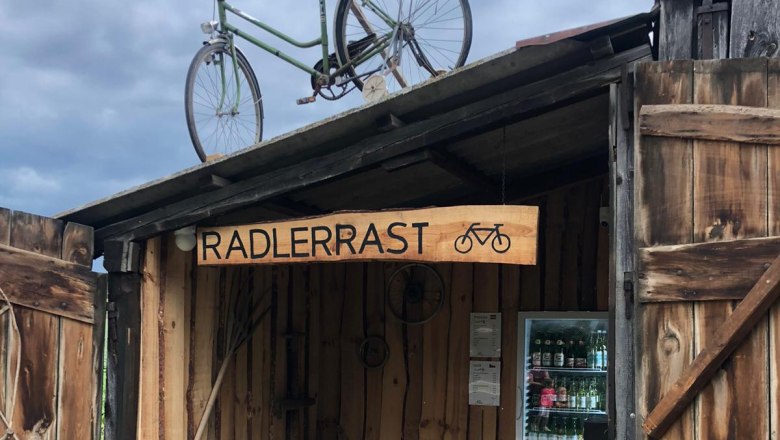 A wooden shelter with a bicycle on the roof and a fridge with drinks. A sign with the inscription 'Radlerrast'.
