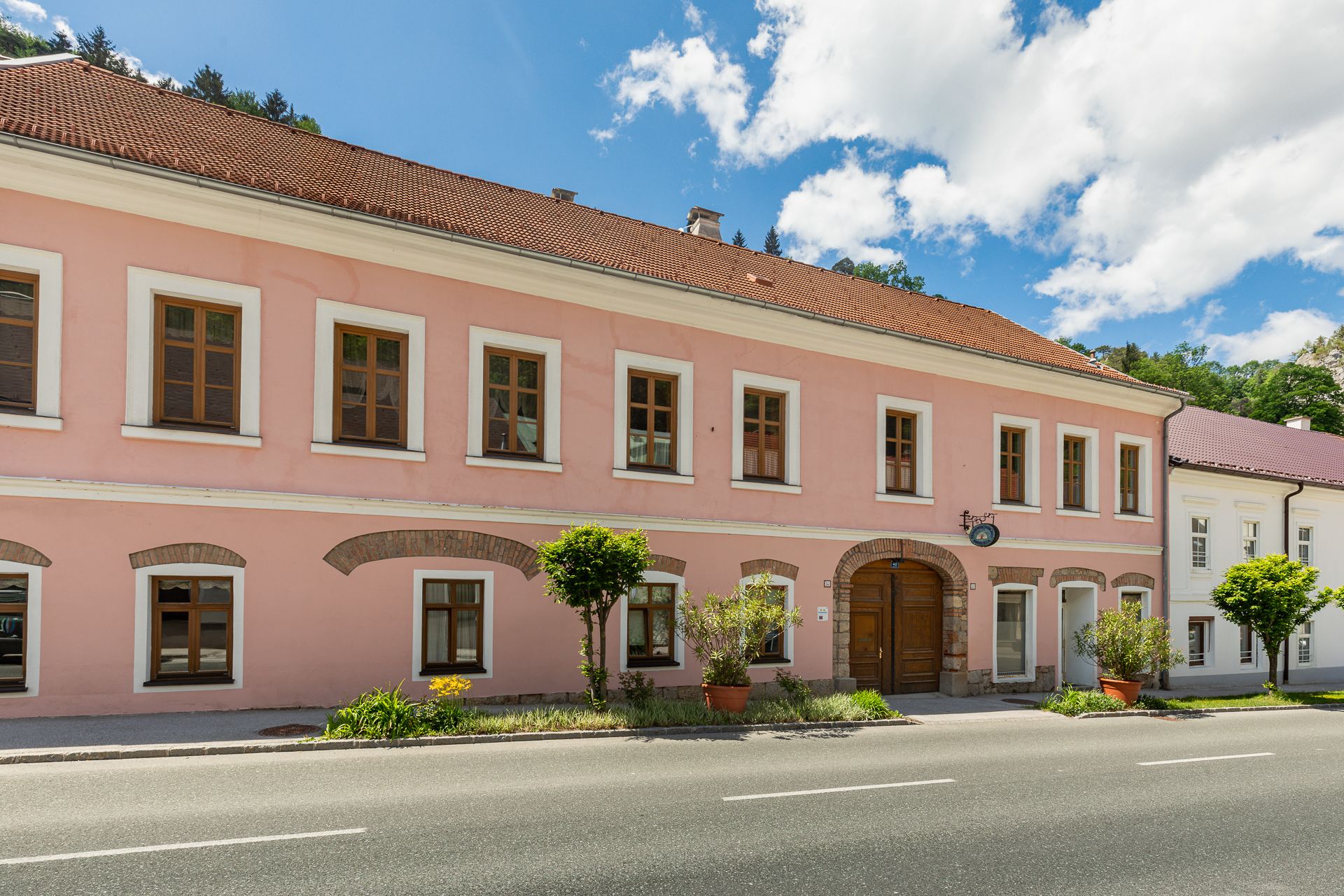 Two-story pink building with brown windows and wooden door, surrounded by plants, under a blue sky.