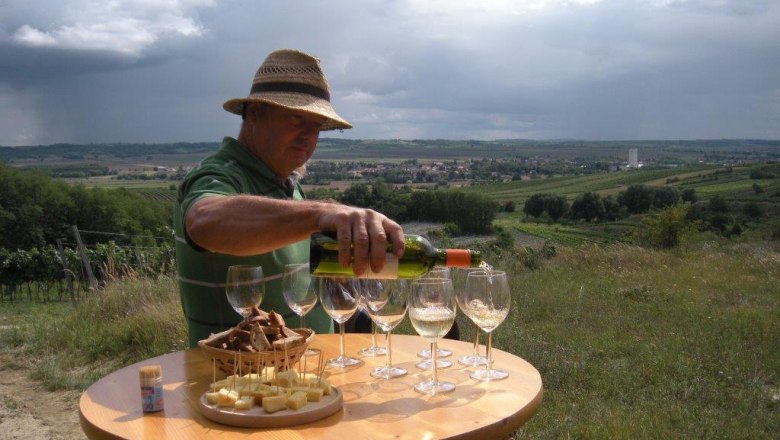 Man pouring wine into glasses on an outdoor table with a view of vineyards and village in the background.