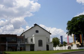 A modern concert hall with white walls and large windows, surrounded by flags and trees, under a cloudy sky.
