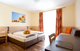 A bright guest room with two beds, a table, a sofa and a picture of sunflowers on the wall.