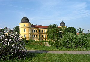 Yellow castle with two towers and a red roof, surrounded by trees and flowers.