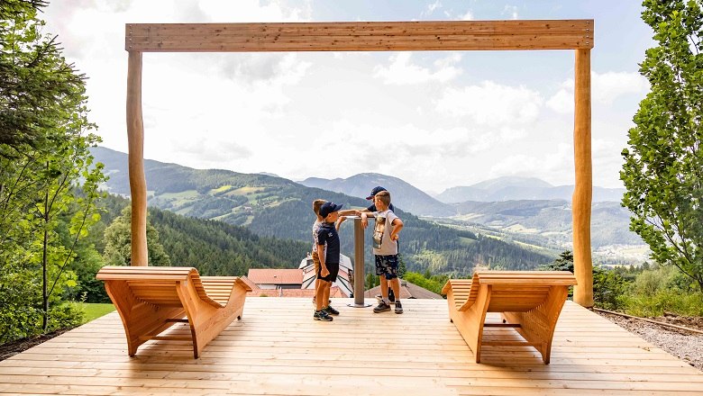 Two children stand on a wooden platform with a view of a mountain landscape.