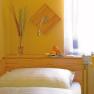 Cozy room with yellow walls, bed, vase with grasses and fruit bowl on a wooden shelf.
