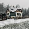 A snow-covered house in traditional style with green shutters, surrounded by trees and a snow-covered street.