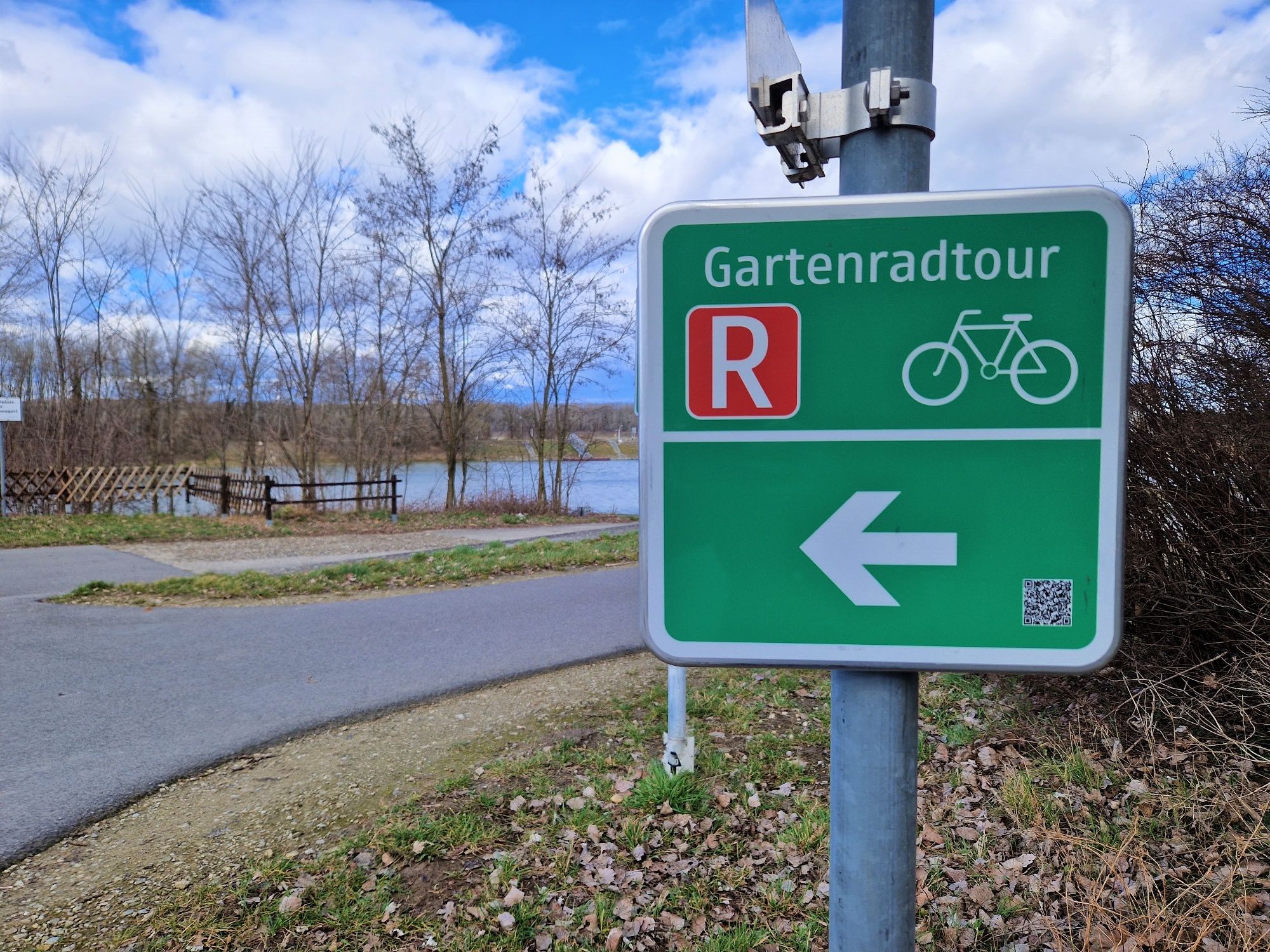 A green sign with the words 'Gartenradtour' and a bicycle symbol points to the left. A path and a lake can be seen in the background.