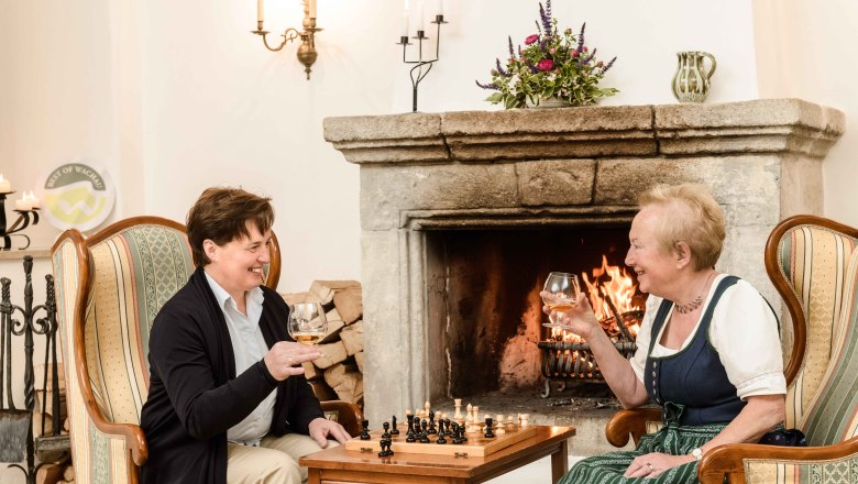 Two women are sitting in armchairs in front of a fireplace, drinking wine and playing chess.