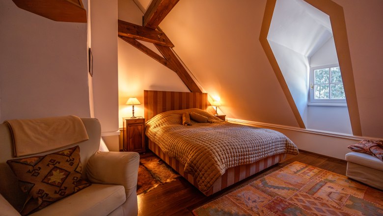 Cozy bedroom with wooden beams, large bed, armchair and carpet. Warm light from bedside lamps, window with view to the outside.