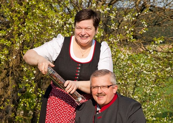 Country inn Heinrichsberg, © Marcella Wallner A man and a woman in traditional dress smile outdoors, the woman holding a large pepper mill.