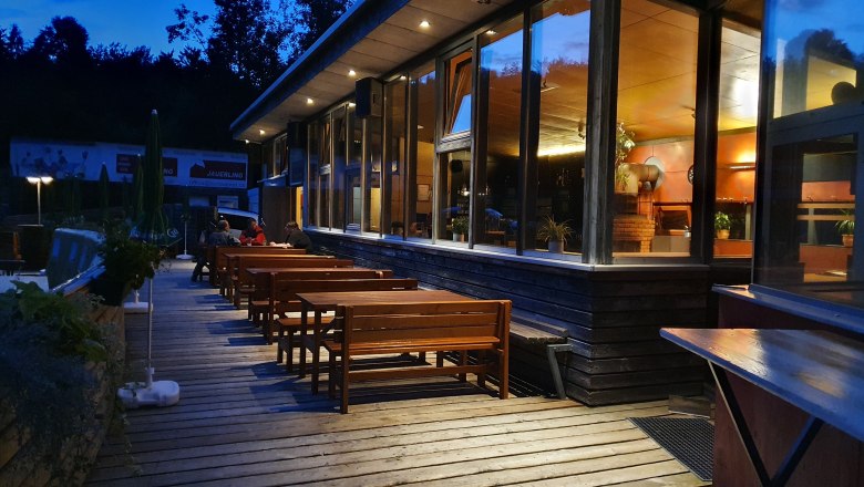 Evening atmosphere at a hut with a wooden terrace and benches, illuminated by warm light, surrounded by trees.