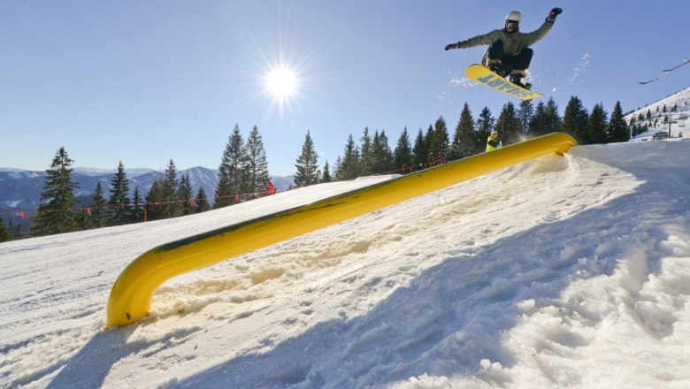 Snowboarder jumping over a yellow obstacle in the Gemeindealpe snowpark in the sunshine.