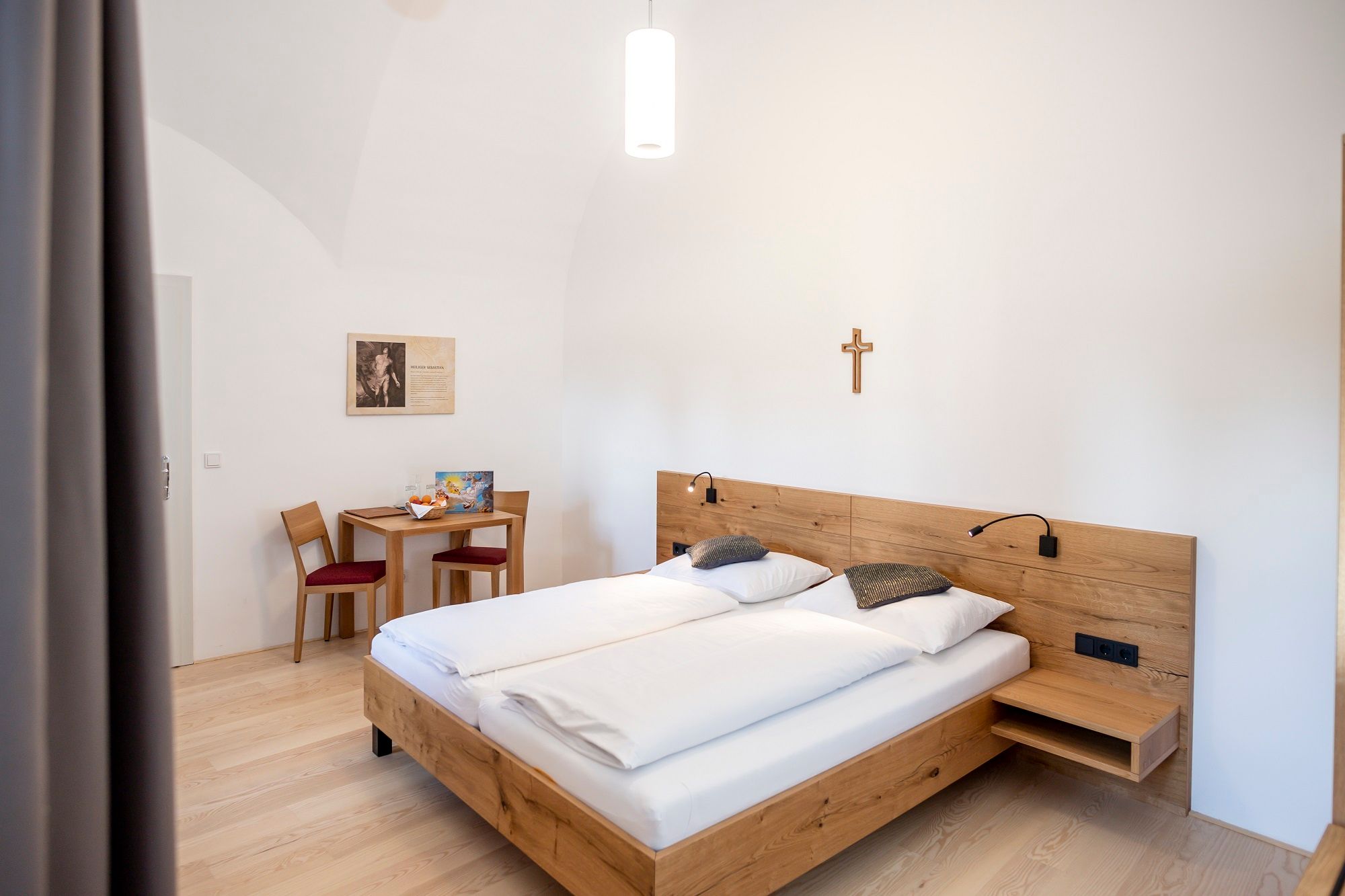 A simply furnished guest room with a double bed, wooden furniture and a cross on the wall.