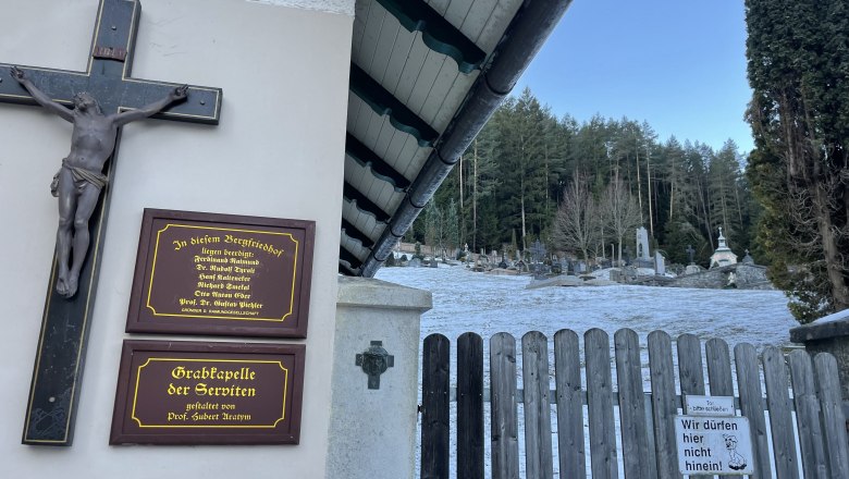 Entrance to the Gutenstein mountain cemetery with wooden gate, cross and memorial plaques. Graves and trees can be seen in the background.