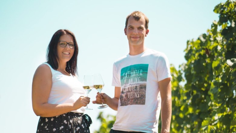 Two people clink glasses outdoors, surrounded by vines.