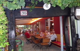 People sit at wooden tables in a covered outdoor area with vines.
