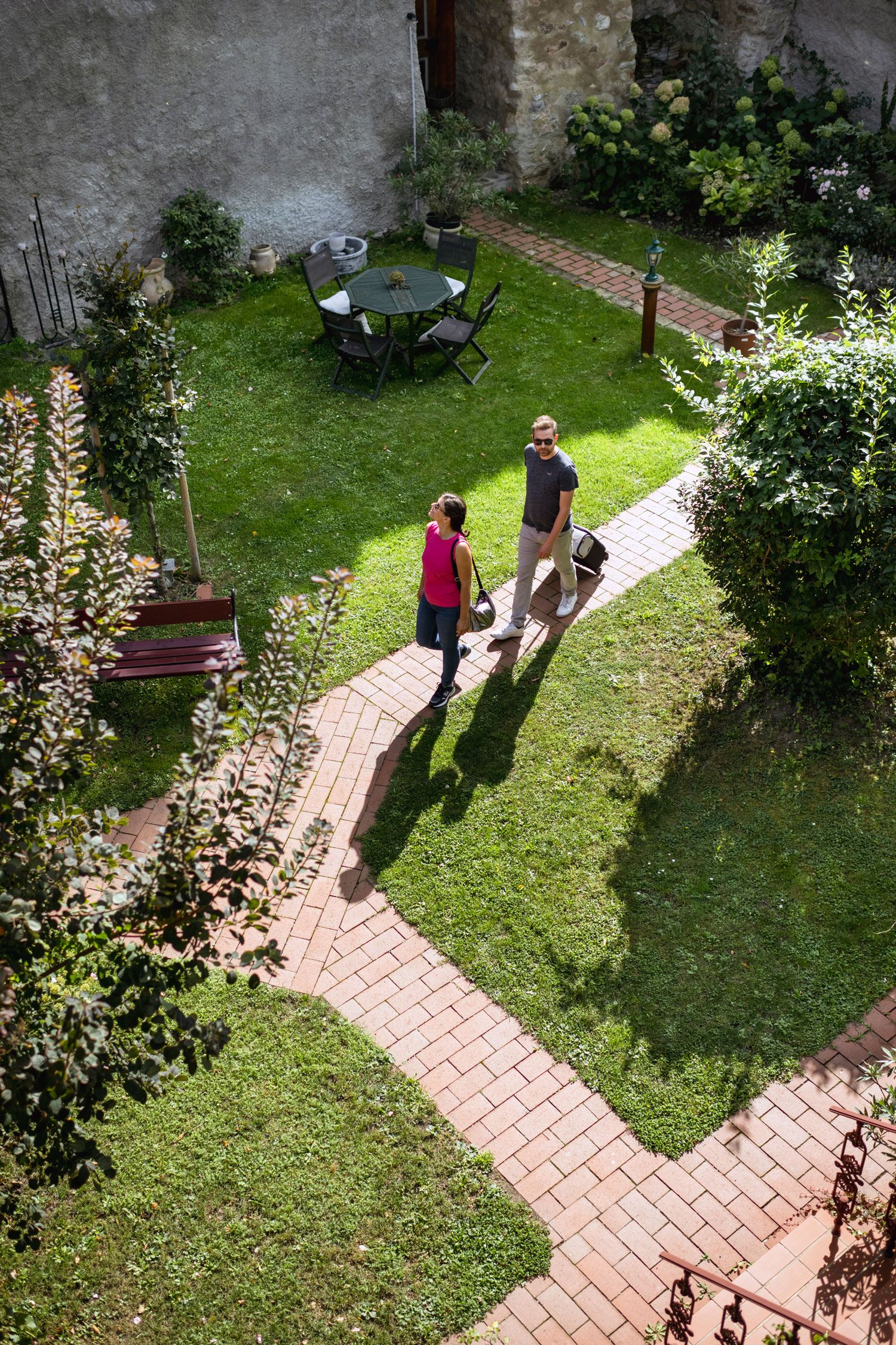 Two people are walking on a paved path through a green garden with a table and chairs.