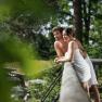A couple in towels lean smiling against a railing outside, surrounded by green trees.