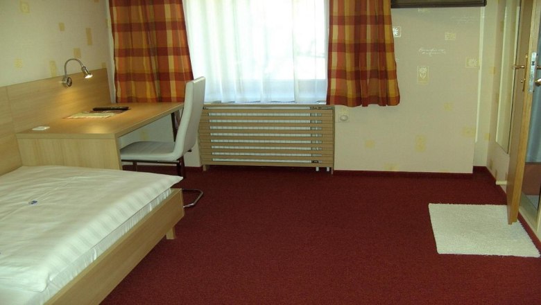 A simply furnished hotel room with a single bed, desk and chair. Red carpet and checkered curtains.