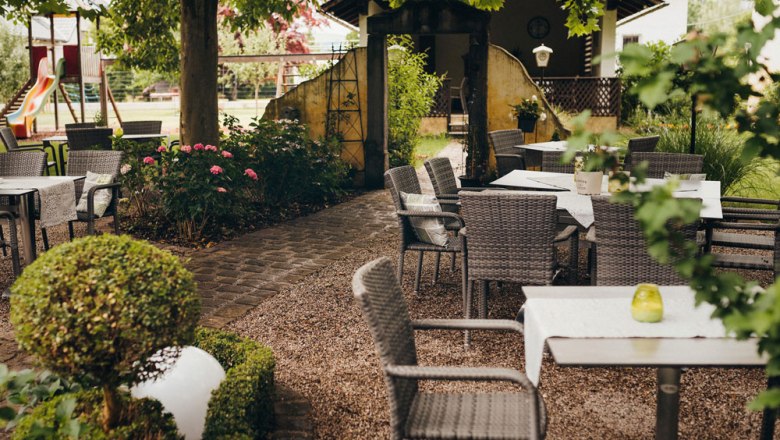A cozy outdoor area with tables and chairs under trees, surrounded by plants and a playground in the background.