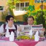 A man and a woman are sitting at an outdoor table set with glasses and cutlery. The man is reading a newspaper.