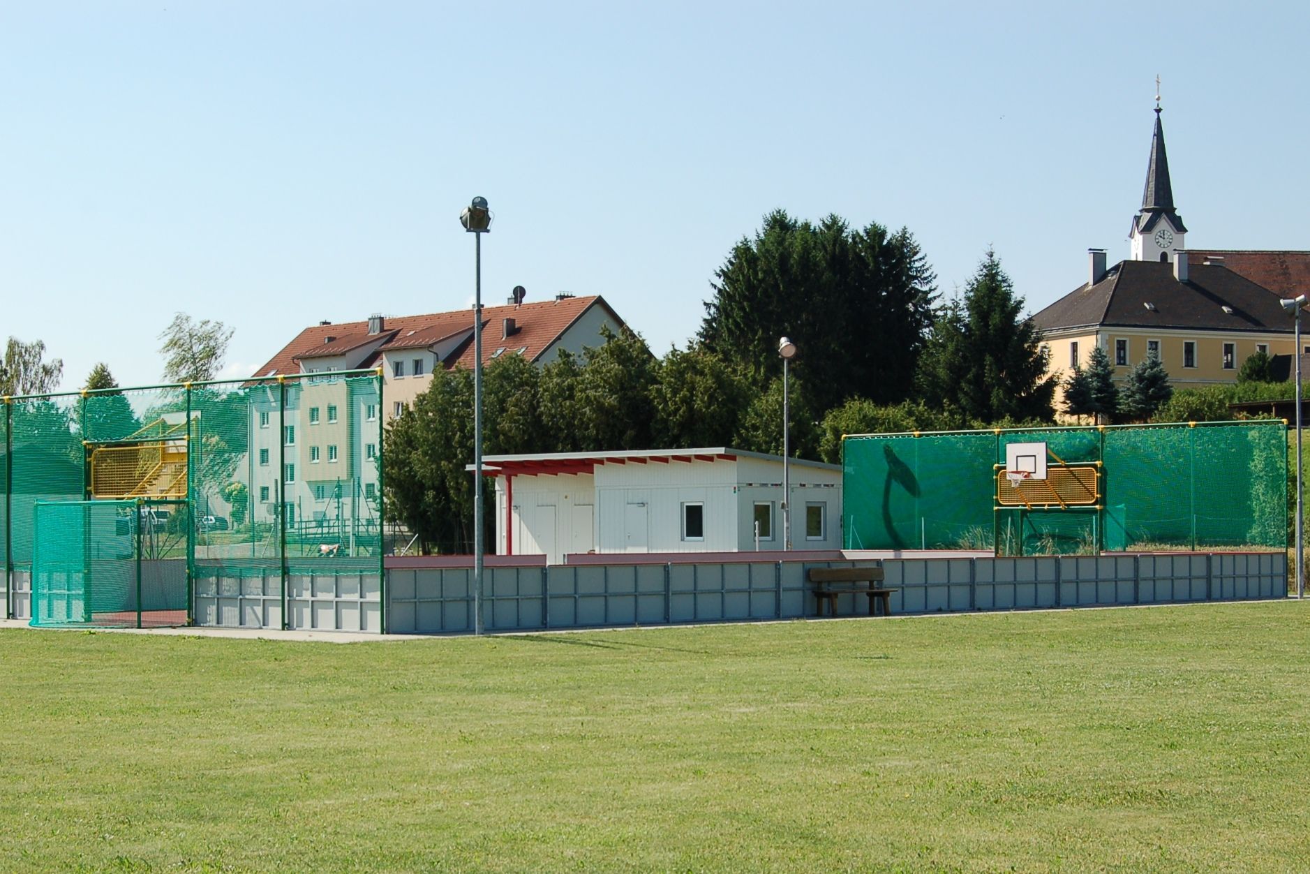 A fun court with basketball hoop and green net, with a small building next to it. Residential buildings and a church can be seen in the background.