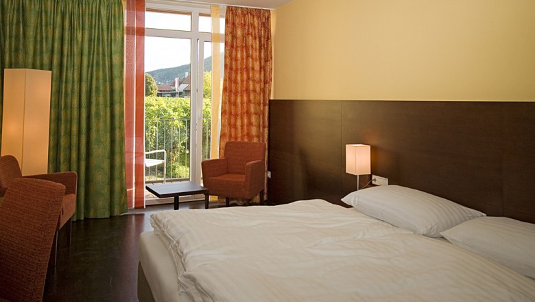 Hotel room with double bed, two armchairs, curtains and balcony with a view of the green landscape.