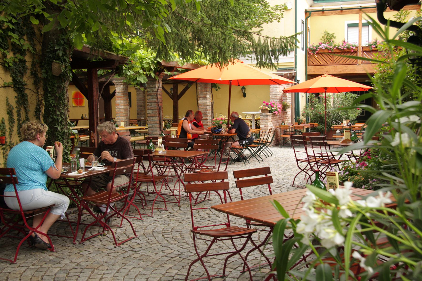 A cozy beer garden with wooden tables, red chairs and orange parasols. People sit and eat outdoors.