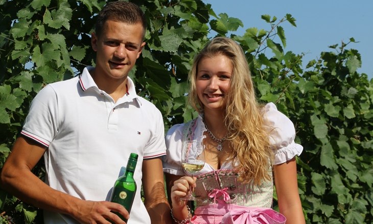Two people are standing in a vineyard, one holding a wine bottle, the other a wine glass. A sign with the inscription 'Settle down' is visible.
