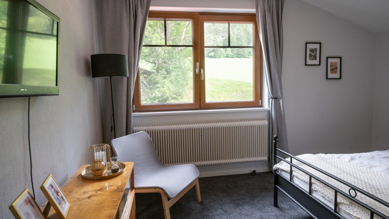Bedroom with gray carpet, bed, armchair, TV and window with a view of the greenery.