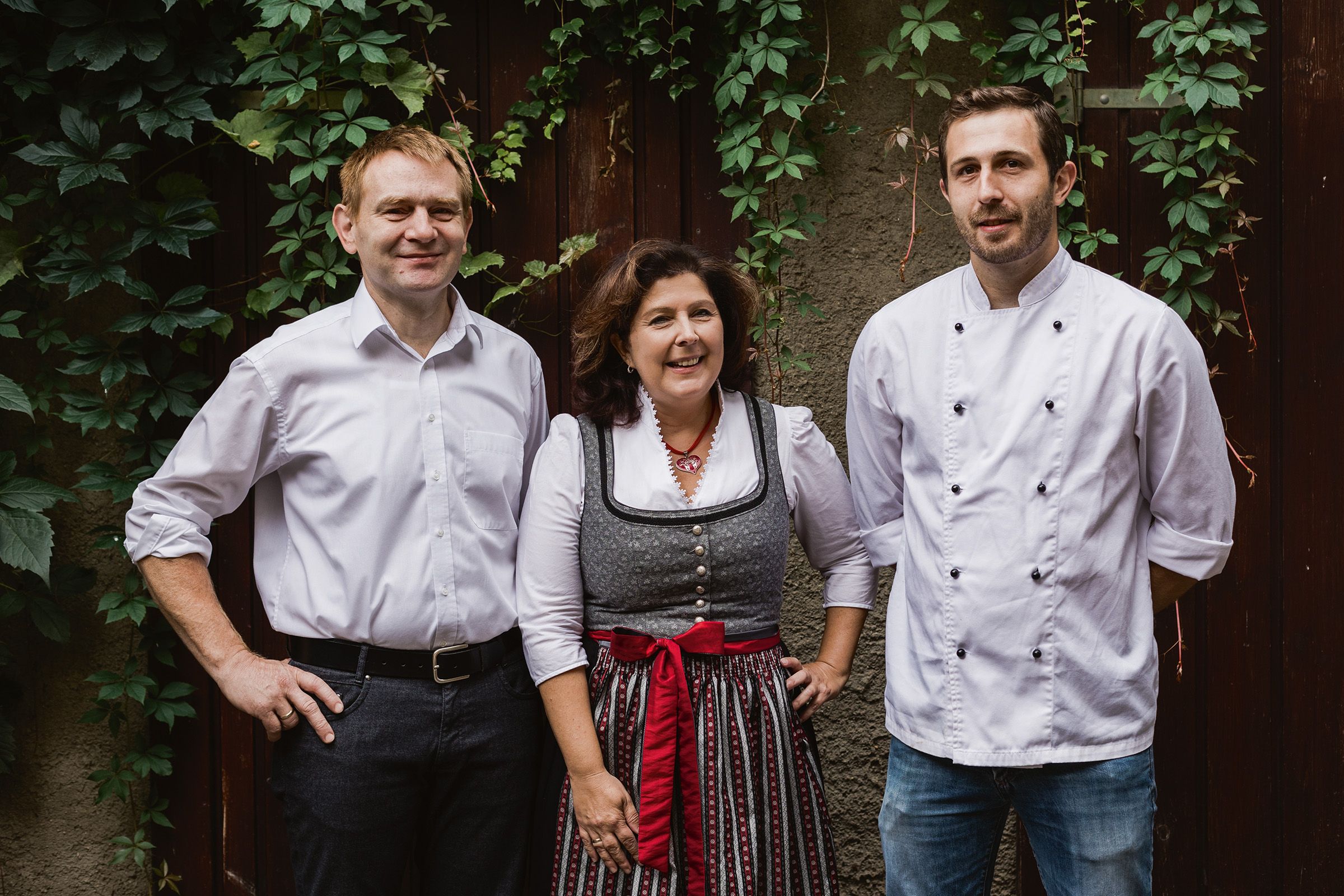 Three people are standing in front of an ivy-covered wall, two men in white shirts and a woman in traditional dress.