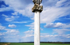A statue on a pillar in a rural landscape under a blue sky with clouds.