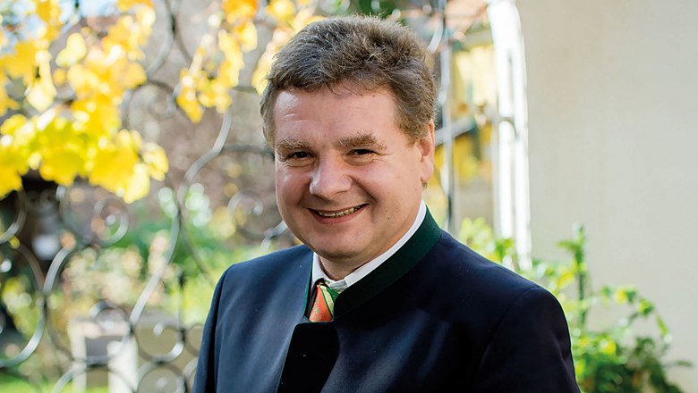 A smiling man in traditional dress stands outside in front of a wrought-iron fence with yellow leaves in the background.