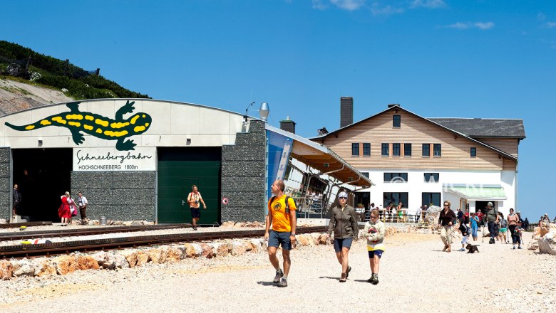 Entrance to the Schneebergbahn on the Hochschneeberg with people and mountain lodge in the background.