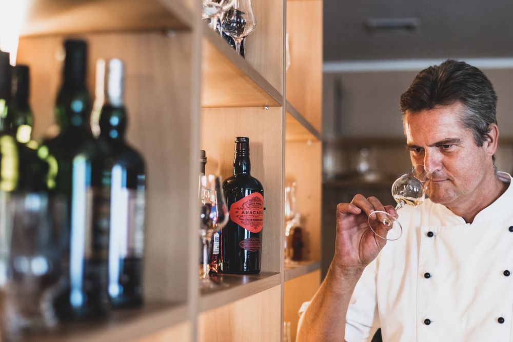 A man in a chef's jacket smells a glass of wine in front of a shelf of bottles.