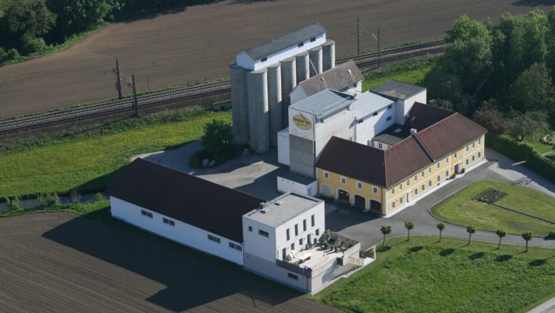 Aerial view of the Rosenfellner mill with buildings and silos, surrounded by fields and railroad tracks.