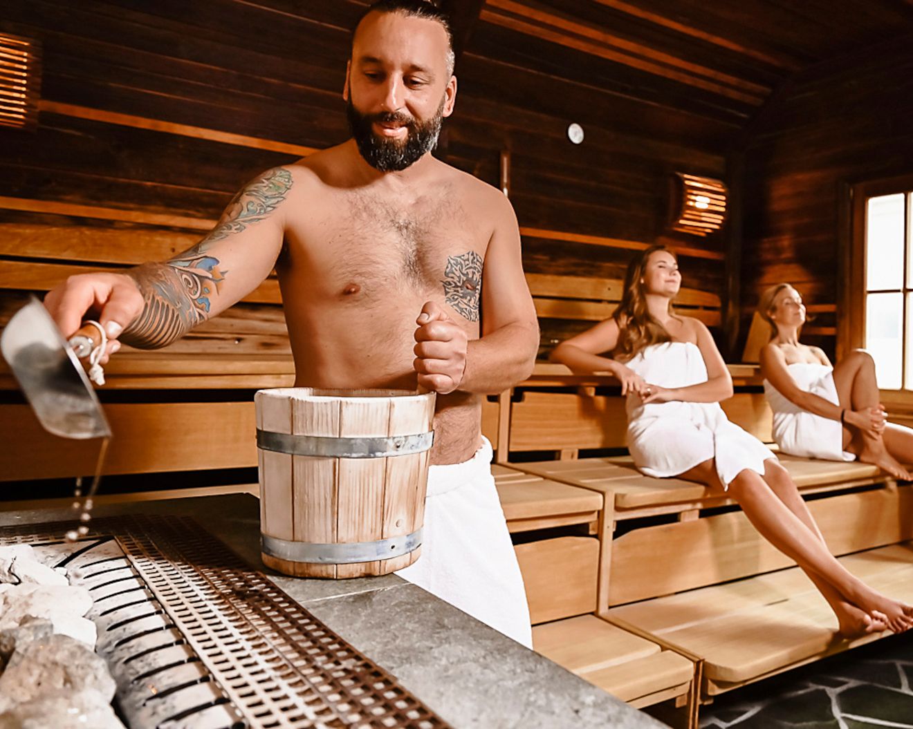 Man pouring water into sauna, two women sitting relaxed.